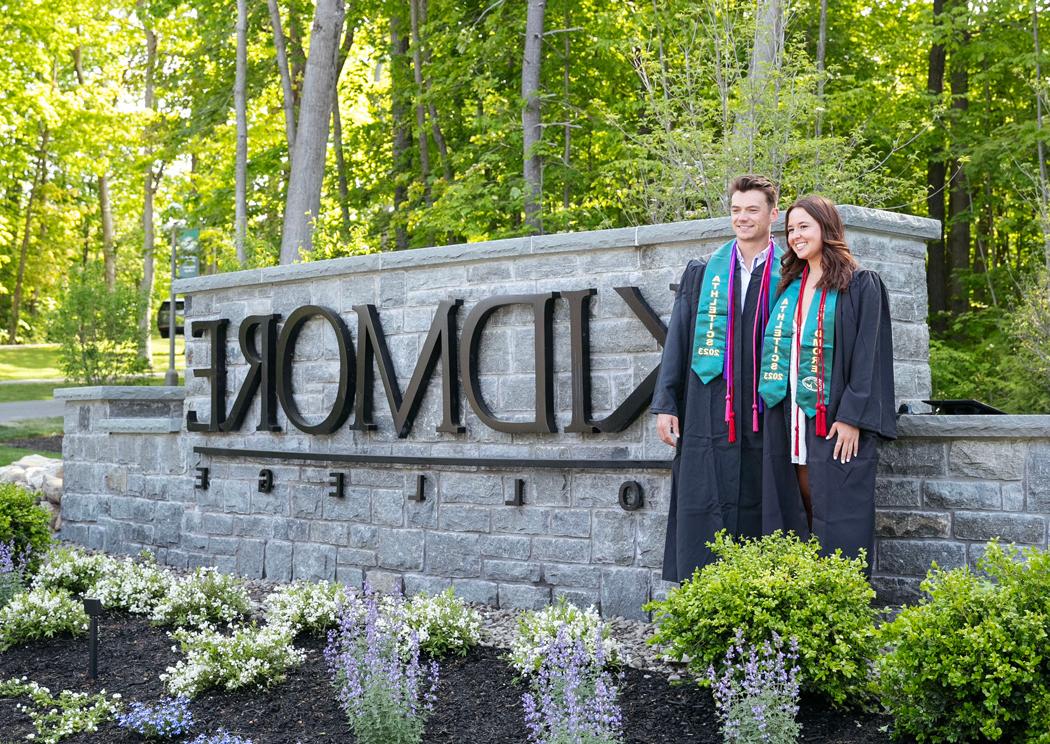 two college graduates post in front of a campus entrance sign that says Skidmore College